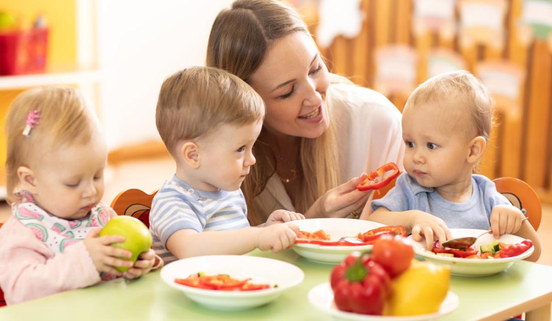 Eine Erzieherin sitzt mit drei kleinen Kindern beim gemeinsamen Essen in einer Kindertagesstätte. (c) Oksana Kuzmina / AdobeStock