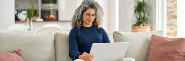 Eine Frau sitzt im Schneidersitz auf einem Sofa mit dem Laptop auf dem Schoß