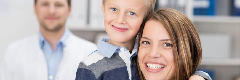 Mutter und Sohn in der Zahnarztpraxis. Sie lächeln in die Kamera, im Hintergrund der Kieferorthopäde