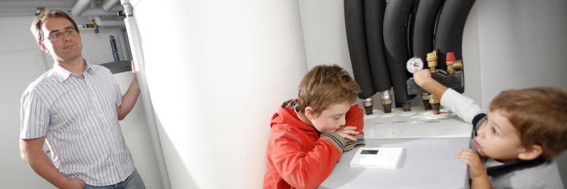 Ein Vater steht im Keller seines Hauses neben seiner Wärmepumpe. Zwei Kinder sind auch anwesend.