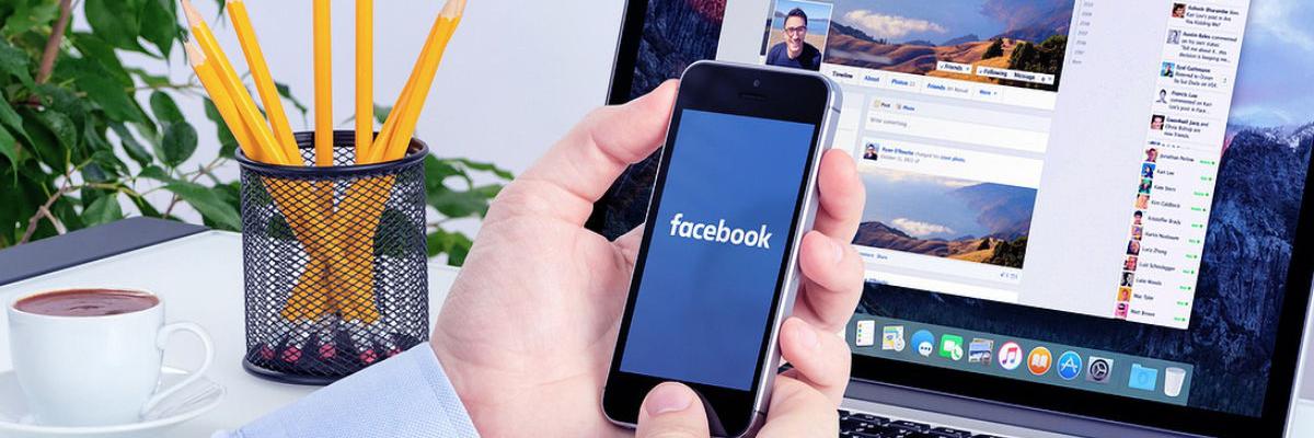 Mann am Schreibtisch hält Smartphone mit dem Schriftzug facebook auf dem Display in seinen Händen, dahinter ein aufgeklappter Laptop mit geöffneter Facebook-Seite
