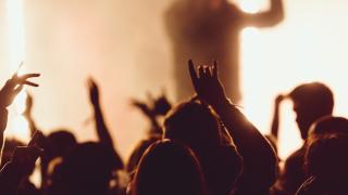 Eine Konzerthalle mit Fans, die die Hände hochrecken. Im Hintergrund steht der Künstler auf der Bühne.