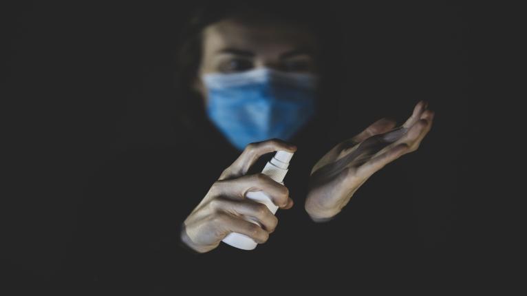 Frau mit Gesichtsmaske sprüht sich Hände mit Desinfektionsmittel ein.